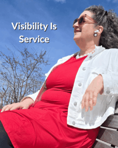 Andrea Stenberg is sitting on a park bench wearing a red dress and white jean jacket. She has on sunglasses as she looks into the distance.
